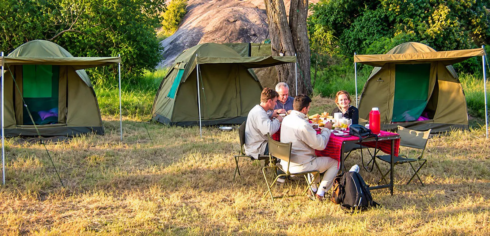 Safari guests sharing a meal beside canvas tents at a bush campsite